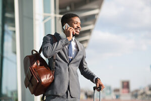 Business Travel. Black Businessman Having Phone Conversation Standing With Suitcase Near Airport